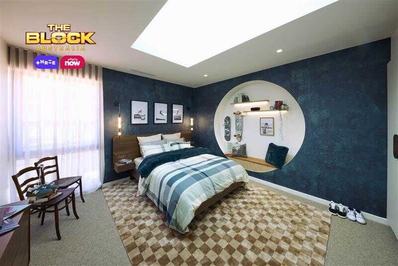 Blue bedroom with a double bed, textured blue wallpaper and blue checked linen. The bed is on a brown checked rug and there is a circular recess in the wall with a bench seat and clothing hooks.