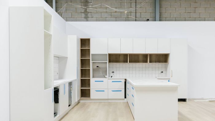 Modern white kitchen under installation with flat-panel cabinets, open wood shelving, a large island bench, tiled backsplash, light timber flooring, and exposed wiring visible above unfinished walls.