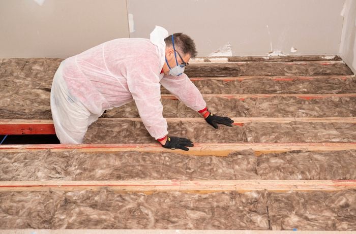 A person wearing protective gear is laying insulation batts between floor joists in a room under construction. 