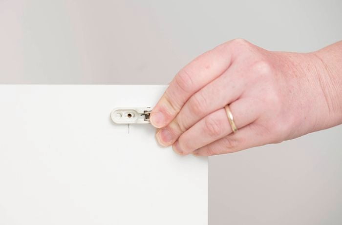 A person is holding a small cabinet hinge bracket against the edge of a cabinet door with circular hinge holes that have already been pre-drilled
