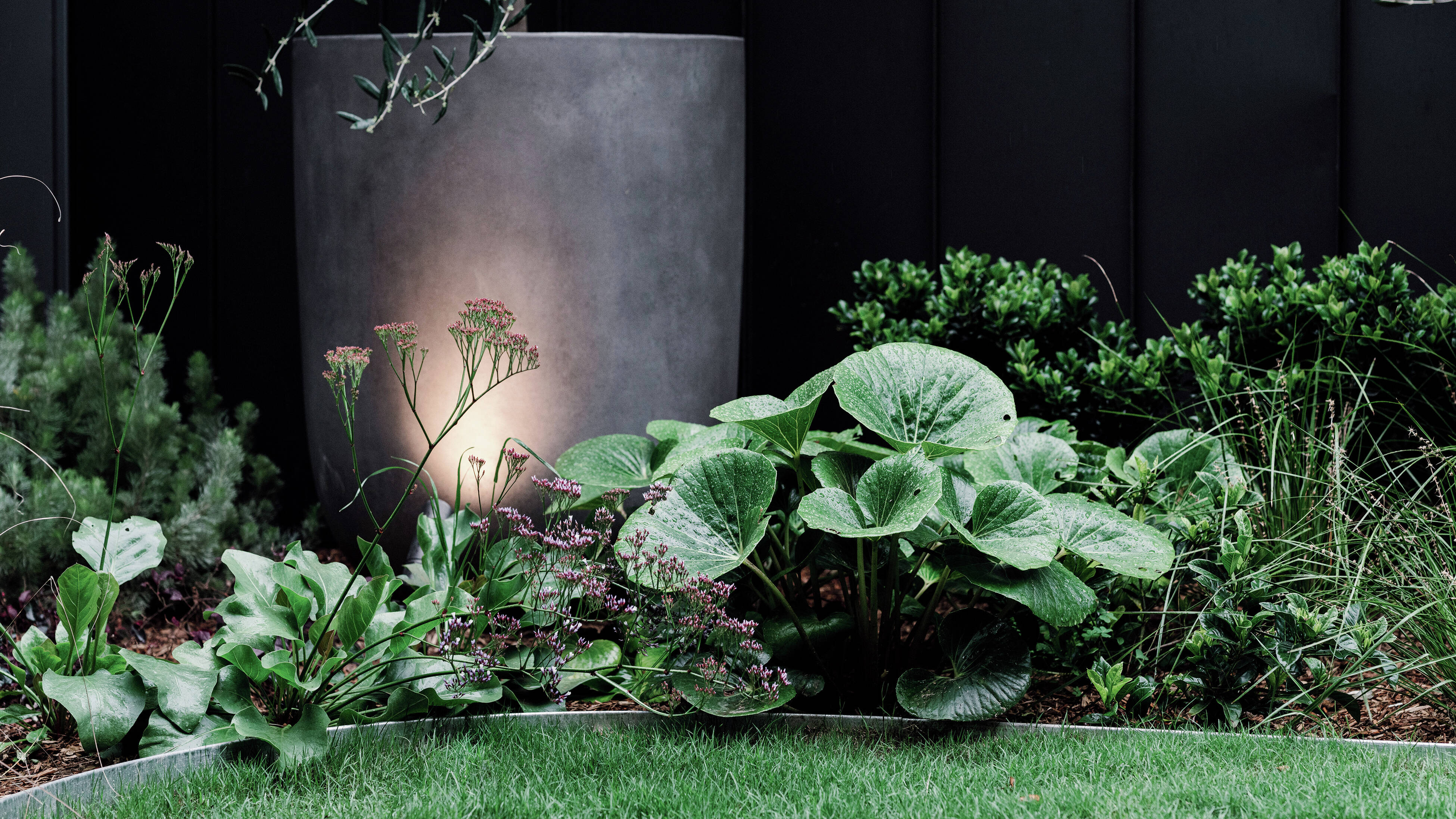  A garden bed with a large grey planter, green foliage, and pink flowers, illuminated by an upward-facing light.]