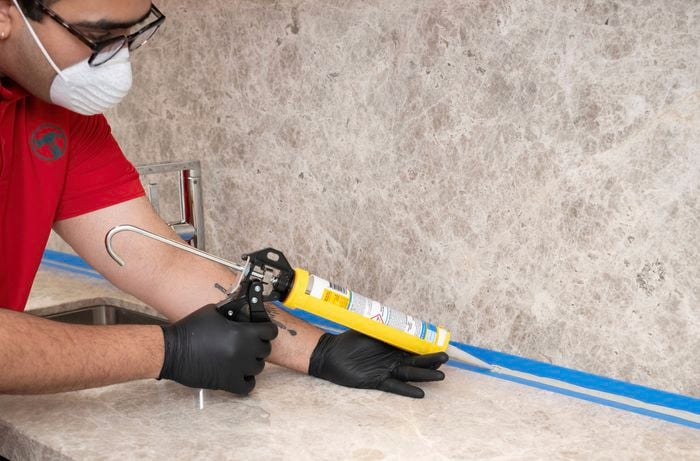 Person wearing protective gloves, glasses, and a dust mask applies a bead of silicone along the joint between a stone-look benchtop and splashback using a caulking gun, guided by blue masking tape.