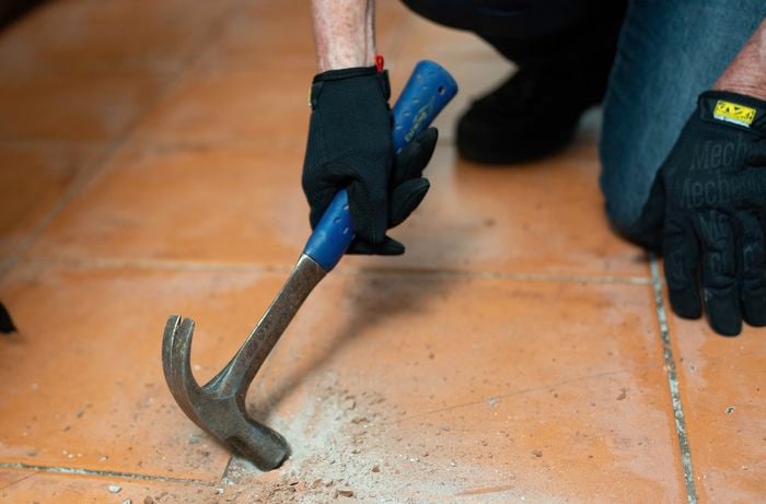 A person wearing gloves uses a claw hammer to break tiles on a dusty floor.