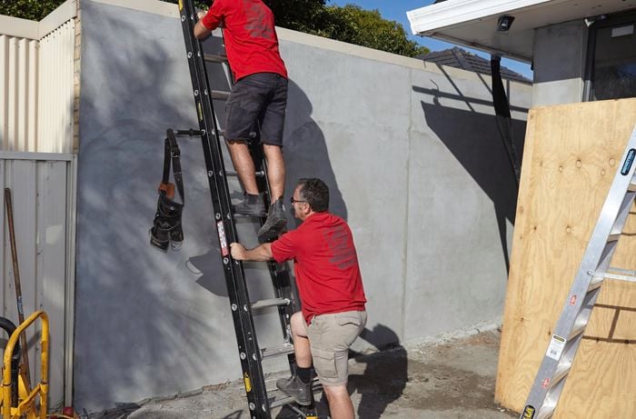 One man holding an extension ladder against a wall while another man climbs