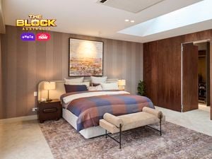 A spacious, modern bedroom with a large bed dressed in a plaid blanket and pillows. The room includes a skylight, a wooden accent wall, a built-in fireplace with a seating area, and large windows with curtains that allow natural light to enter.