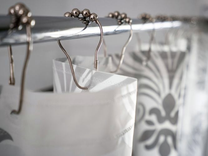 Close up of a shower curtain rod with decorative hooks holding a grey fleur-de-lis patterned curtain with white background