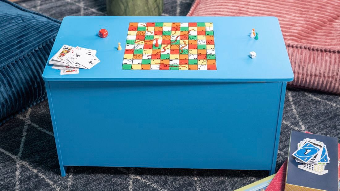 A finished DIY games storage box painted blue with a snakes and ladders board on the lid, styled with dice, playing cards and game pieces.