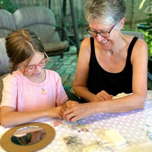 Child and grandmother smiling while they create mosaics at a table together. They are sitting in a patio setting.