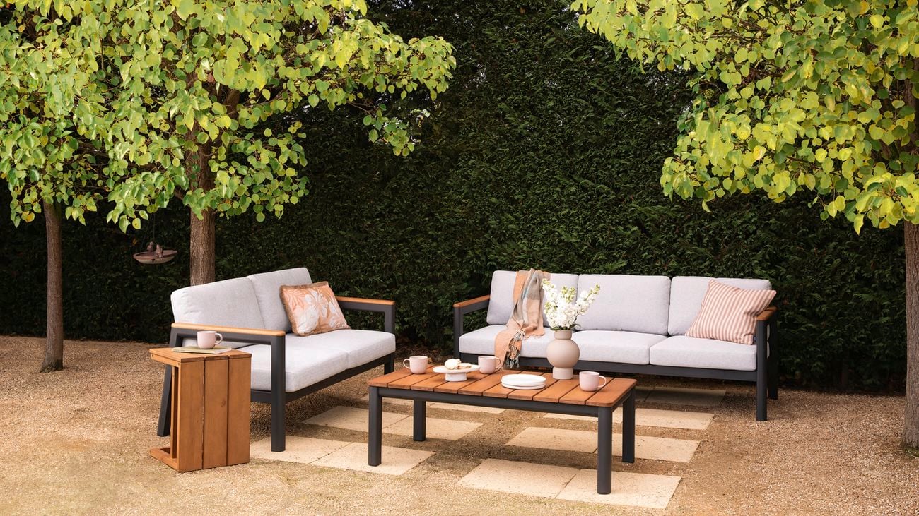 An open-air patio with an outdoor furniture setting, and large green trees and a hedge creating shade.