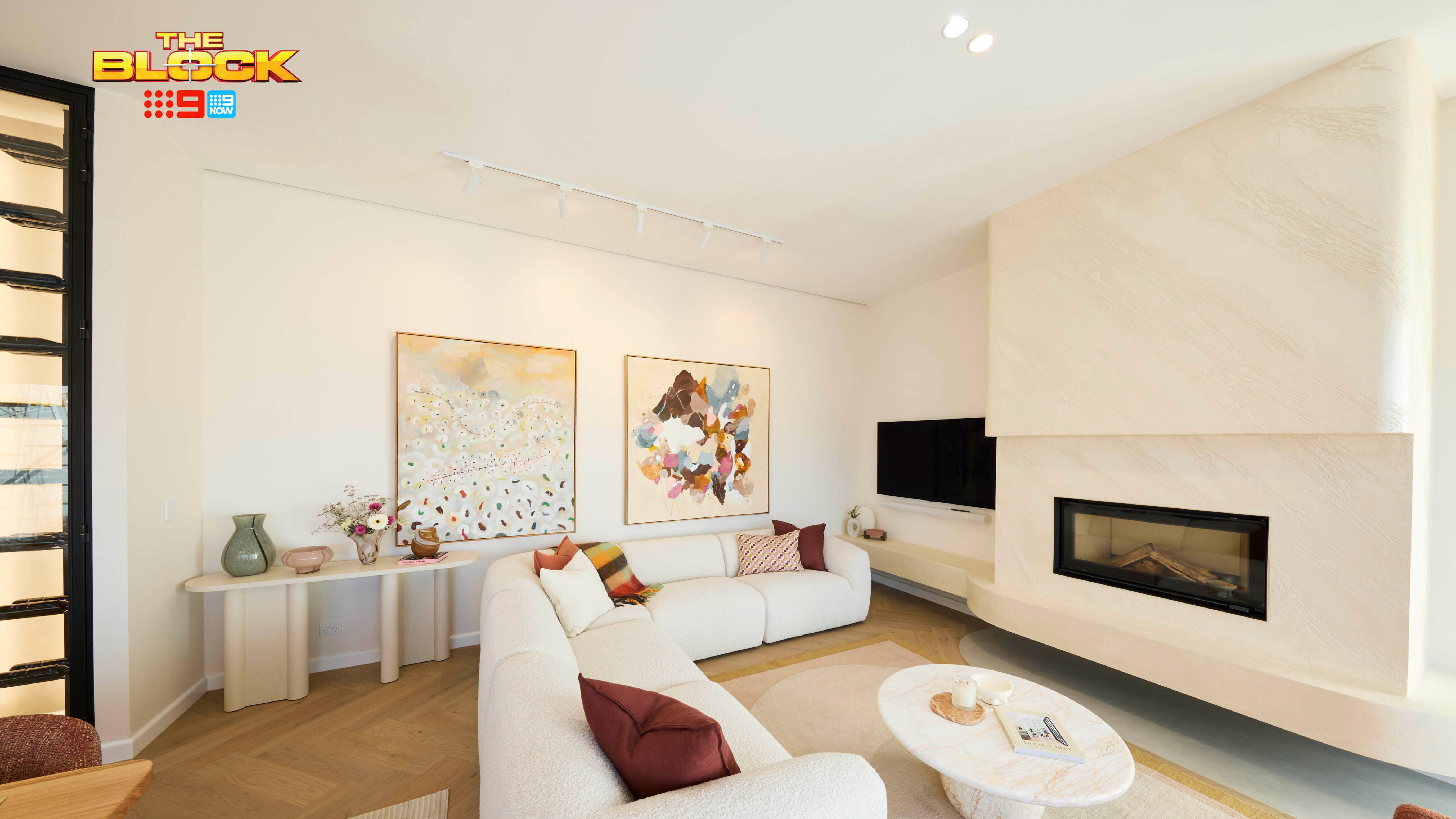 A modern living room featuring a white sectional sofa, a round coffee table, and a built-in fireplace. Two large abstract paintings hang above a console table with decorative items. The room has light-colored walls and wooden flooring. 