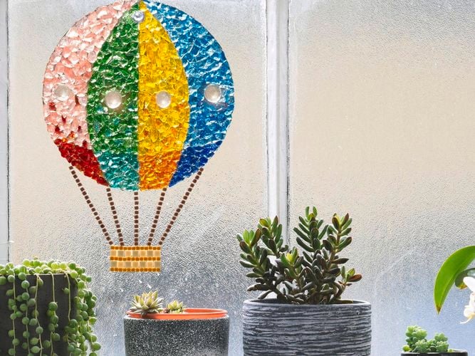 Brightly coloured mosaic Hot Air Balloon sun catcher attached to a window. There are plants on the window sill. 