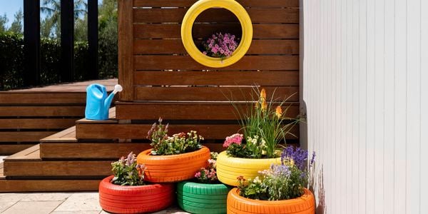 An outdoor patio styled with brightly painted tyre planters overflowing with flowers, arranged beside a timber deck and complemented by a hanging yellow tyre planter on the fence. 
