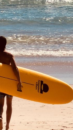 Surfer carrying a yellow Summer vibes longboard under one arm towards the waves.