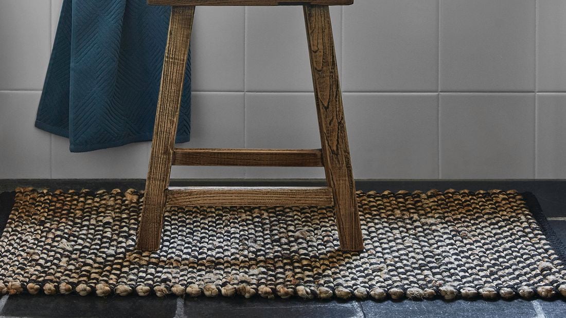 A wooden stool sitting on a rug against a pale grey tiled wall