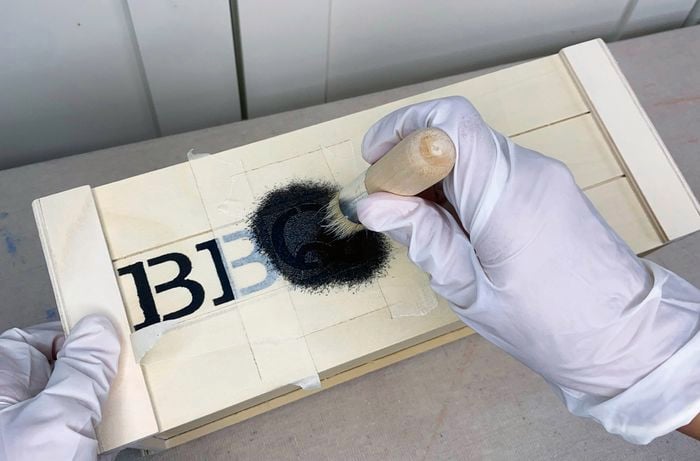 Gloved hands dab black paint onto stencil letters on the side of a small timber crate.