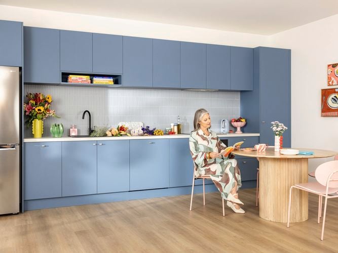 Woman in early 50's reading a book at a timber table. A straight line blue kitchen is in the background with floral arrangement on the bench.