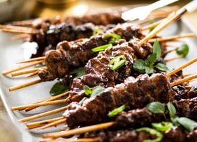 A white platter piled with BBQ skewers garnished with sliced green chilli and oregano leaves.