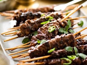 A white platter piled with BBQ skewers garnished with sliced green chilli and oregano leaves.