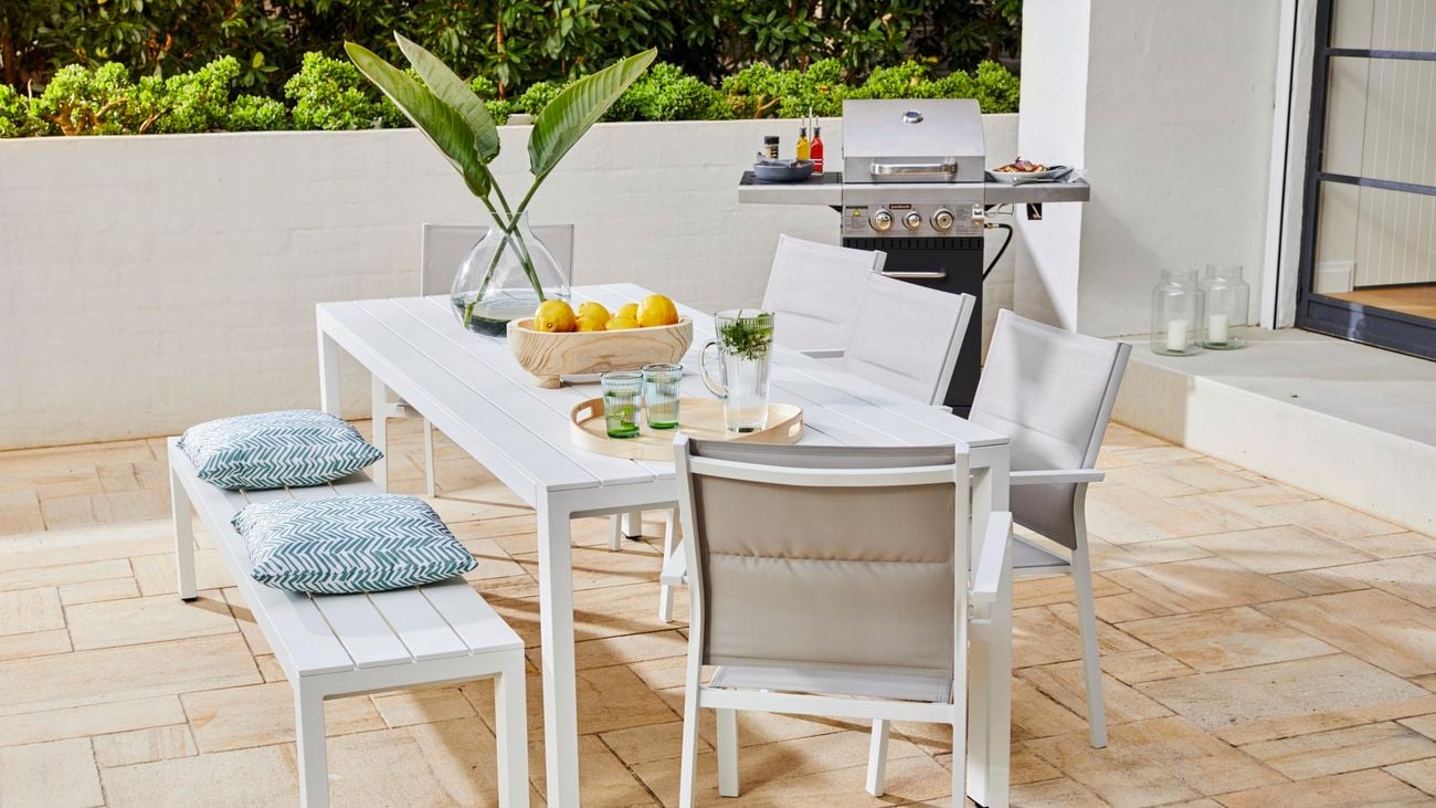 White aluminium outdoor dining table on a patio featuring a barbecue and bench seat