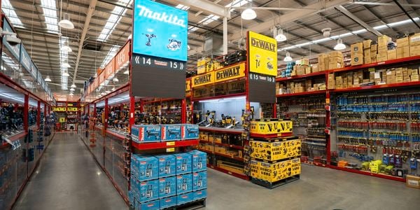 Wide view of a Bunnings power tools department featuring Makita and DeWalt displays. Red shelving units hold boxed tools and accessories, while large overhead brand signs mark the aisles beneath a high industrial ceiling with bright lighting.