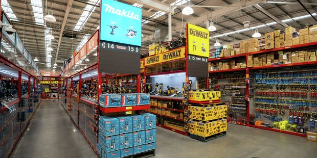 Wide view of a Bunnings power tools department featuring Makita and DeWalt displays. Red shelving units hold boxed tools and accessories, while large overhead brand signs mark the aisles beneath a high industrial ceiling with bright lighting.