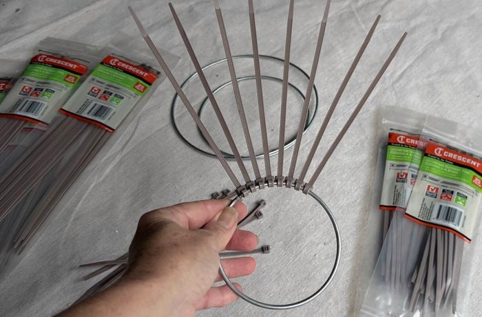 Close-up of a hand attaching grey plastic cable ties evenly around a circular metal ring, with unopened packs of cable ties placed on both sides on a light fabric surface.