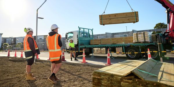Two workers in high-vis clothing watching as a crane lifts a timber pack from a delivery truck at a construction site.