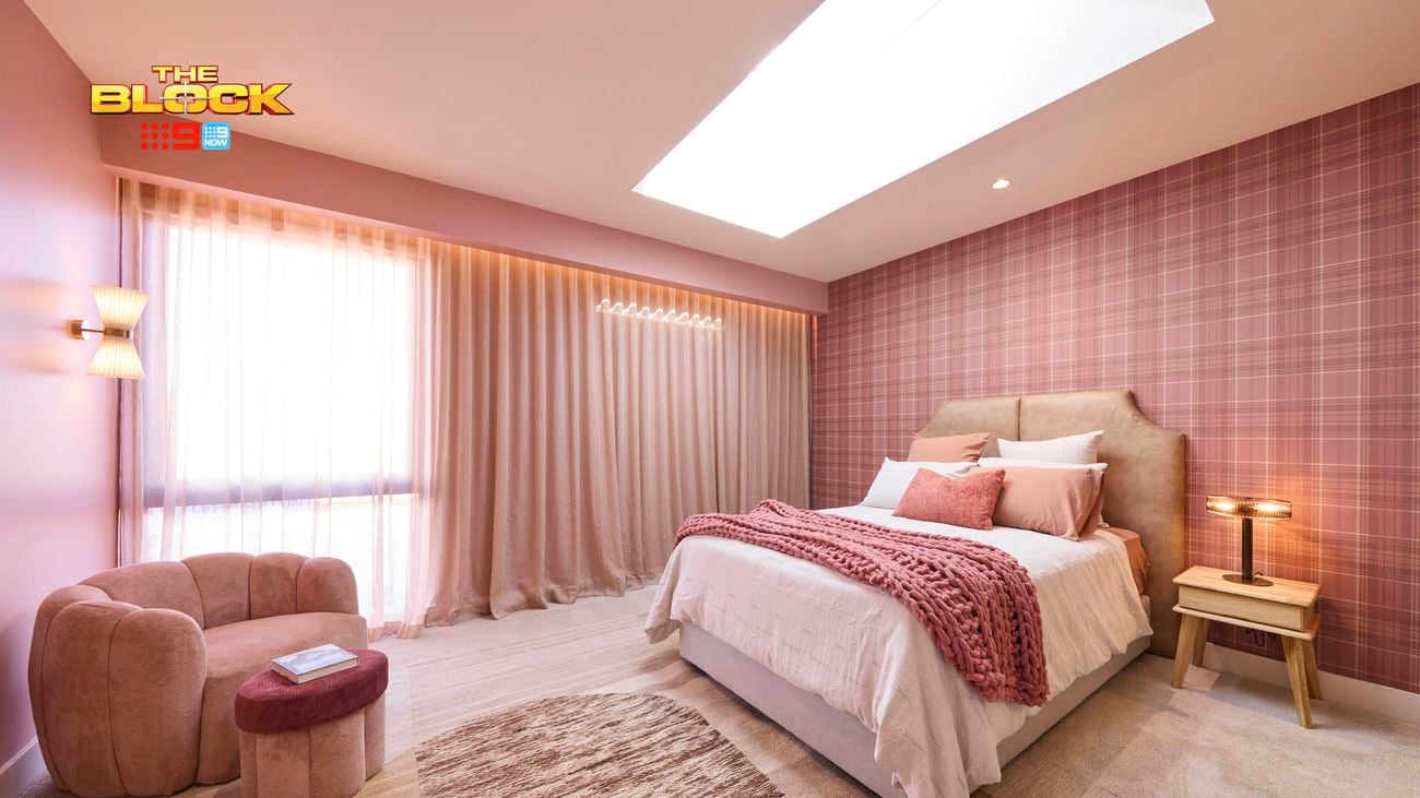 Pink bedroom with a check wallpapered wall and a double bed with pink linens. Opposite is a large velvet chair and matching foot stool.