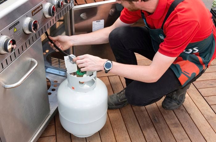 A person crouching in front of a BBQ checking the hose to the gas bottle