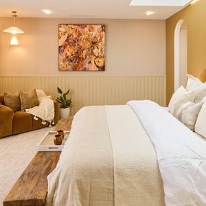 A stylish bedroom featuring a large bed with multiple pillows and a beige color palette. A wooden bench sits at the foot of the bed. To the left, there’s a cozy seating area with a brown sofa, cushions, and two small tables. Large windows with sheer white curtains let in natural light. The walls are decorated with artwork and lighting fixtures. 