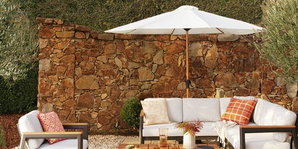 Outdoor patio lounge setting featuring a white-cushioned sectional sofa and matching armchair around a wooden coffee table on white gravel. A large white umbrella shades the seating area, with a rustic stone wall, manicured hedge, and olive trees creating a landscaped garden backdrop. Decorative cushions, a vase of flowers, and drinks on the table add a warm, inviting atmosphere.