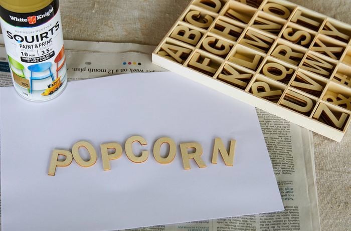 A piece of paper with wooden craft letters spelling ‘popcorn’ laid out on top. A can of spray paint and a box set of craft letters are next to it.