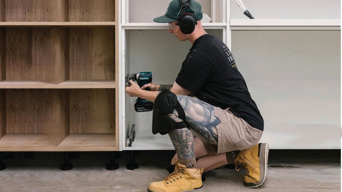A tradesperson wearing a cap, earmuffs, and safety glasses kneels, using a power drill to install a white cabinet next to wooden shelving