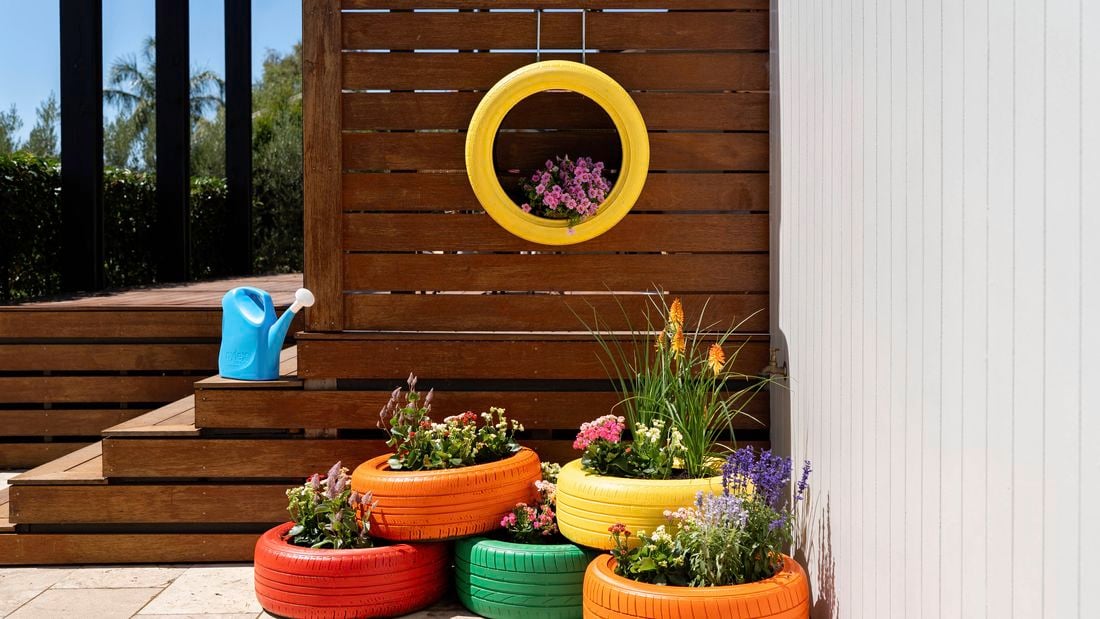 An outdoor patio styled with brightly painted tyre planters overflowing with flowers, arranged beside a timber deck and complemented by a hanging yellow tyre planter on the fence. 