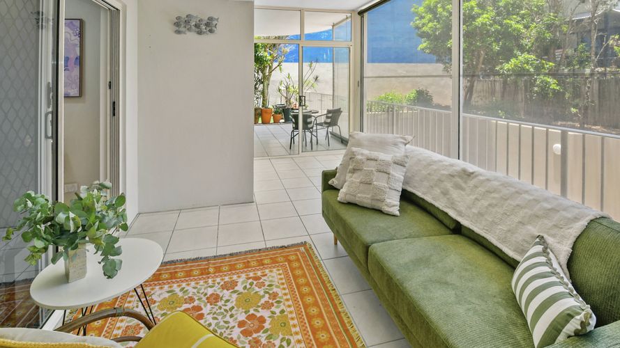 Light-filled enclosed patio or sunroom with a green sofa, patterned rug, small side table and screened windows overlooking leafy gardens. Glass doors open to an outdoor seating area.