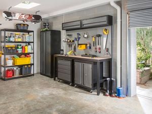 Organized garage with metal shelving, a snowboard mounted on the ceiling, black cabinet, workbench with drawers, wall-mounted cabinets, pegboard with hanging tools, and boots near an open garage door