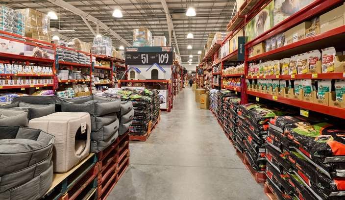 A wide aisle inside a Bunnings store with tall red metal shelving units line both sides, stocked with bags of pet food, treats, and other supplies. On the left are stacks of plush pet beds and larger pet accessories, while the right side displays rows of packaged pet food. Overhead industrial lighting illuminates the concrete floor and high metal ceiling. A sign toward the back shows pricing for a large dog kennel.