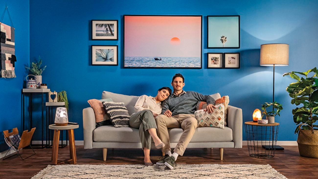 A couple sitting on a couch in a living room with blue walls, picture frames on the wall, lamps and indoor plants.