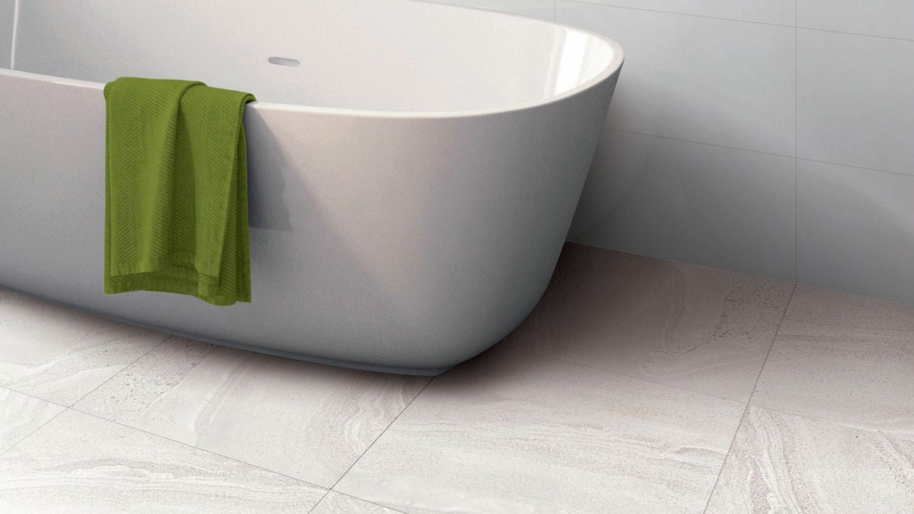 A white freestanding bathtub with a green towel draped over its side, set against light-coloured tiled floor and walls.