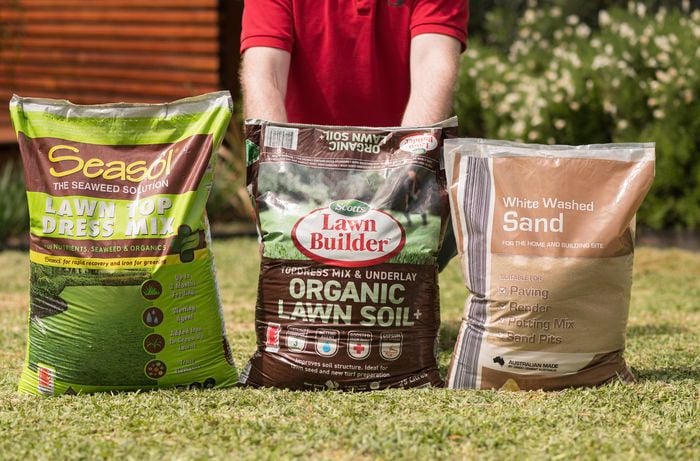 Three lawn care product bags, including topdressing mix, organic lawn soil, and white washed sand, are arranged on a lawn to choose the right topdressing mix