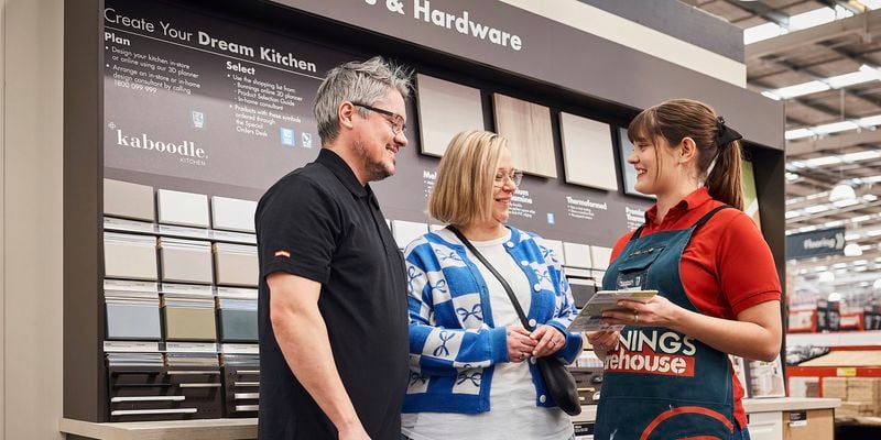 Store team member in a Bunnings Warehouse apron shows kitchen cabinet samples to two customers standing beside a wall display of cabinet finishes at a Kaboodle kitchen planning station.