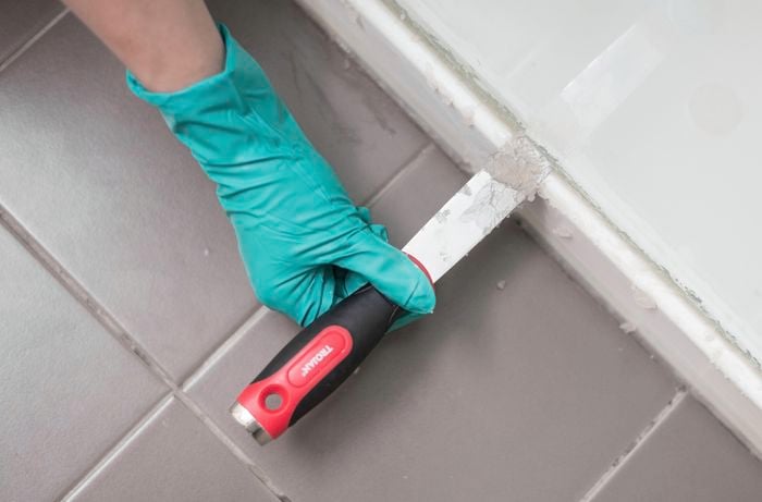 A hand wearing green gloves is using a Trojan paint scraper to apply silicone caulk remover