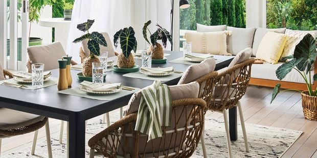 A bright indoor–outdoor dining area featuring a black dining table set with glassware, neutral table settings, and small decorative plants. Woven chairs with light cushions surround the table. To the right is a cosy lounge area with cream and yellow cushions on a wooden sofa. Large windows and greenery outside create an airy, natural atmosp