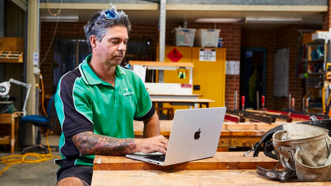 A tradesperson in a green and black polo shirt sits at a wooden workbench using a silver laptop, with tools and workshop equipment visible in the background.]