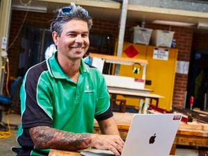 A smiling man in a green and black polo shirt with tattoos is working on a laptop in a workshop setting.]