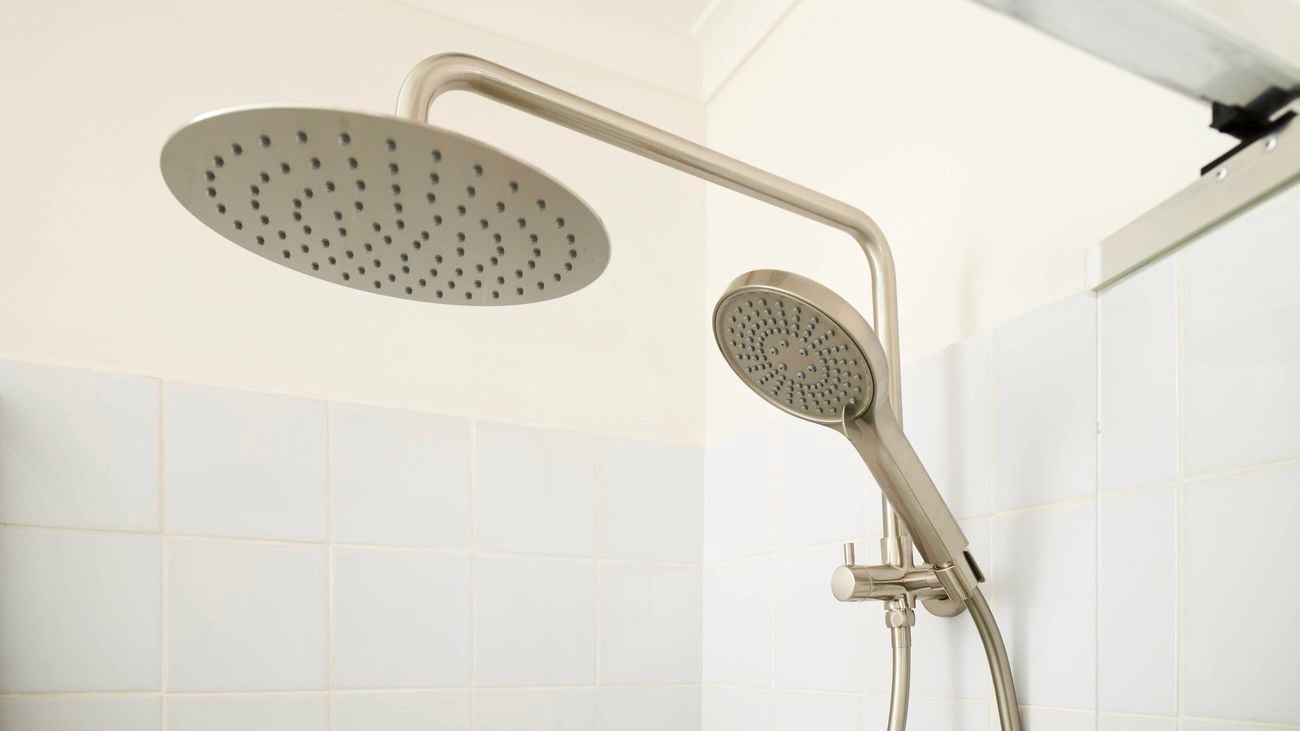 Close-up of a dual shower setup with a large round overhead rain shower and a handheld showerhead on a vertical bar, both in a brushed metal finish, set against white tiled walls in a modern bathroom.