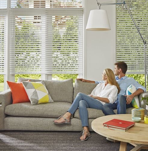 Young couple sitting in a modern lounge room looking out a window with white venetian blinds