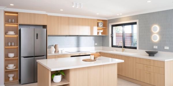 A bright, modern kitchen with natural sunlight features light wood cabinetry, a large island with white countertops, and a window