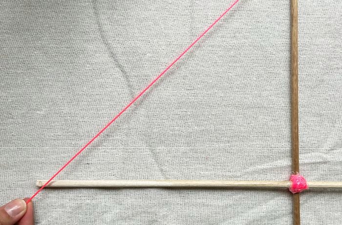 Two wooden dowels forming a cross, joined with pink tape, with a neon pink string running diagonally on a grey fabric background.