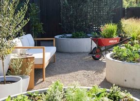 Exterior of a black timber modern farmhouse with an outdoor area featuring a red wheelbarrow, raised garden beds, potted plants, and a white outdoor sofa on gravel. Trees, lawn and greenery are visible in the background.